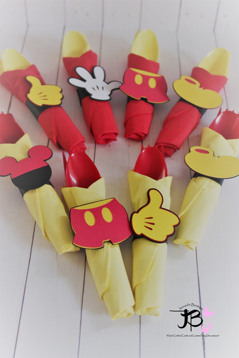 May include: Arrangement of red and yellow napkins, each holding a red spoon. The napkins are decorated with Mickey Mouse-themed cutouts, including gloves, shorts, and ears. Ideal for a themed party.