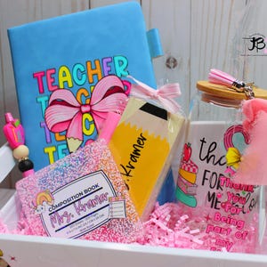 May include: A white gift basket filled with teacher appreciation gifts. The basket includes a blue notebook with the words "Teacher" in rainbow letters, a pink and white composition book with the words "Mrs. Kramer" on the cover, a yellow pencil-shaped bookmark with the name "Kramer" on it, and a clear glass jar with a pink ribbon and the words "Thank You for Being Part of My Crew" on it.