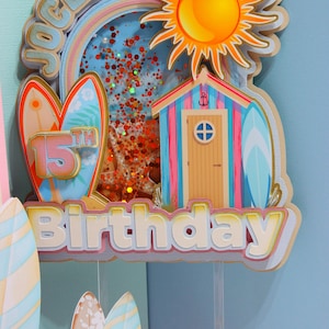 Puede incluir: Adorno de pastel de cumpleaños colorido con temática de playa. Presenta un sol, una cabaña de playa, tablas de surf y las palabras "Jocelyn 15th Birthday". El adorno está decorado con purpurina y un arcoíris.