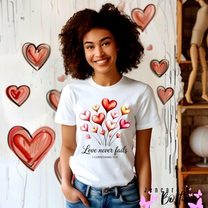 Könnte beinhalten: Ein weißes T-Shirt mit einem Design aus Aquarellherzen in Rot, Rosa und Gelb. Der Text "Love never fails" und "1 Corinthians 13:8" ist unter den Herzen aufgedruckt. Der Hintergrund zeigt gezeichnete Herzen.