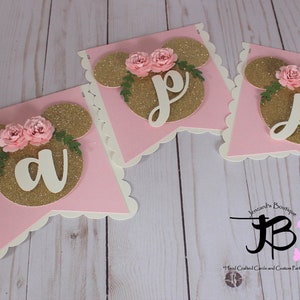 Puede incluir: Bandera de brillo rosa y dorado con la palabra "Happy" escrita en letras blancas. Cada letra est&aacute; decorada con una cabeza de Mickey Mouse de brillo dorado con flores rosas y hojas verdes.