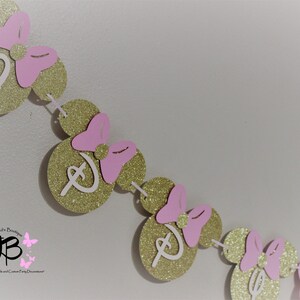 May include: Gold glitter Minnie Mouse ears with pink bows and white letter D's strung together to form a banner.