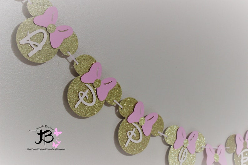 May include: Gold glitter Minnie Mouse ears with pink bows and white letter D's strung together to form a banner.