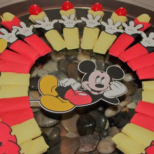 May include: A festive arrangement featuring Mickey Mouse-themed party decorations. The centerpiece includes a Mickey Mouse cutout surrounded by red and yellow napkins, Mickey Mouse hand cutouts, and red plastic spoons. The arrangement is set on a bed of smooth stones.