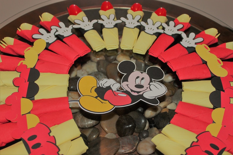 May include: A festive arrangement featuring Mickey Mouse-themed party decorations. The centerpiece includes a Mickey Mouse cutout surrounded by red and yellow napkins, Mickey Mouse hand cutouts, and red plastic spoons. The arrangement is set on a bed of smooth stones.