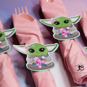 Puede incluir: Cuatro servilletas rosas con anillos de servilleta de personaje verde y marr&oacute;n. El personaje sostiene un ramo de flores rosas. Los anillos de servilleta tienen un borde azul claro. El texto "Simply Beautiful" y un "JB" estilizado son visibles en la servilleta inferior derecha.