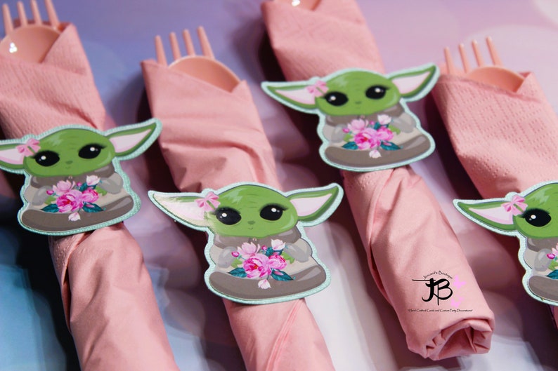 Puede incluir: Cuatro servilletas rosas con anillos de servilleta de personaje verde y marr&oacute;n. El personaje sostiene un ramo de flores rosas. Los anillos de servilleta tienen un borde azul claro. El texto "Simply Beautiful" y un "JB" estilizado son visibles en la servilleta inferior derecha.