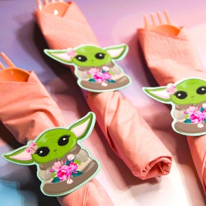 Puede incluir: Tres servilletas rosas con personajes de dibujos animados verdes y marrones que sostienen flores. Los personajes llevan una t&uacute;nica marr&oacute;n y tienen ojos grandes. Las servilletas est&aacute;n envueltas alrededor de tenedores.