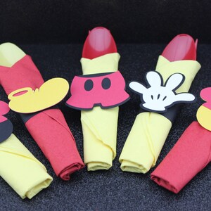 May include: Napkin rings with Mickey Mouse-themed designs. Each napkin is folded in a unique way, with red and yellow napkins. The napkin rings feature Mickey's shorts, gloves, and ears. Perfect for a themed party.