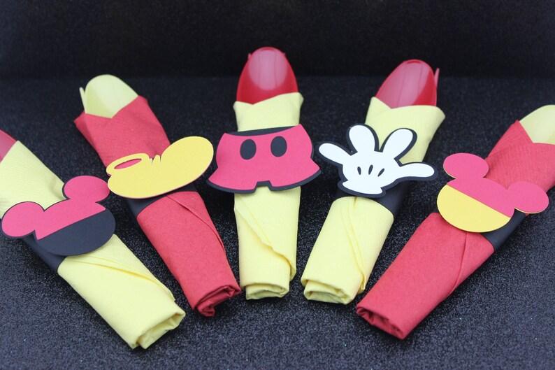 May include: Napkin rings with Mickey Mouse-themed designs. Each napkin is folded in a unique way, with red and yellow napkins. The napkin rings feature Mickey's shorts, gloves, and ears. Perfect for a themed party.