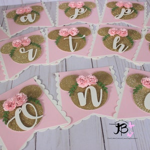 Puede incluir: Una pancarta de brillo rosa y dorado con las letras "Happy" y los n&uacute;meros "One" en blanco. Cada letra y n&uacute;mero est&aacute; en una bandera separada con un fondo rosa y una cabeza de Mickey Mouse de brillo dorado con flores rosas.