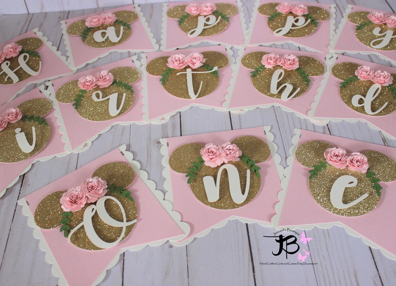 Puede incluir: Una pancarta de brillo rosa y dorado con las letras "Happy" y los n&uacute;meros "One" en blanco. Cada letra y n&uacute;mero est&aacute; en una bandera separada con un fondo rosa y una cabeza de Mickey Mouse de brillo dorado con flores rosas.