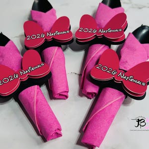 Op de afbeelding: Vier roze servethouders met zwarte keukengerei en roze servetten. Elke servethouder heeft een rode strik met de tekst "2020 Nationals" in het wit. De servetten zijn opgerold en onder de strik geplaatst.