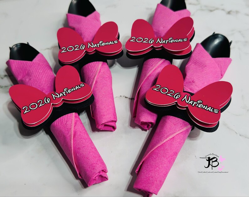 Op de afbeelding: Vier roze servethouders met zwarte keukengerei en roze servetten. Elke servethouder heeft een rode strik met de tekst "2020 Nationals" in het wit. De servetten zijn opgerold en onder de strik geplaatst.