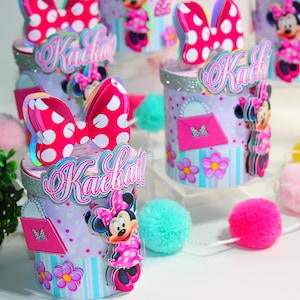 May include: Pink and white Minnie Mouse themed party favor boxes with the name "Kackati" written on them. The boxes are decorated with Minnie Mouse bows, flowers, and a pink purse.
