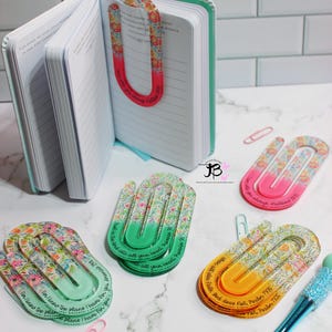 Peut inclure: Une collection d'agrafes à motifs floraux et un journal. Les agrafes ont une forme en U avec des couleurs dégradées et des citations inspirantes. Un stylo avec un manche bleu pailleté est également présent. Le journal est ouvert, affichant des pages lignées.