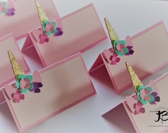 May include: Pink place cards with a unicorn horn and flower embellishments. The horn is gold glitter and the flowers are pink, teal, and purple.