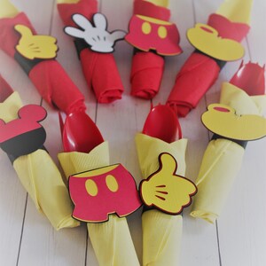 May include: Party decorations featuring red napkins with yellow spoons and napkin rings shaped like Mickey Mouse's gloves, shorts, and ears. The decorations are arranged on a white wooden surface.