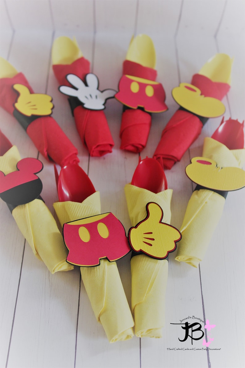 May include: Party decorations featuring red napkins with yellow spoons and napkin rings shaped like Mickey Mouse's gloves, shorts, and ears. The decorations are arranged on a white wooden surface.