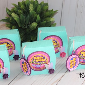 Könnte beinhalten: Fünf türkisfarbene Geschenkboxen mit einem rosa-weißen Hubba Bubba Bubble Tape Logo. Jede Box ist mit einer lila Blume und einer weißen Feder verziert. An jeder Box ist ein lilafarbiges Etikett mit dem Hubba Bubba Logo befestigt.