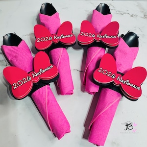 Op de afbeelding: Vier roze servetarrangementen met zwarte keukengerei en rode strikaccenten. Elke strik heeft de tekst "2026 Nationals" in het wit. De servetten zijn opgerold en op een decoratieve manier gerangschikt.