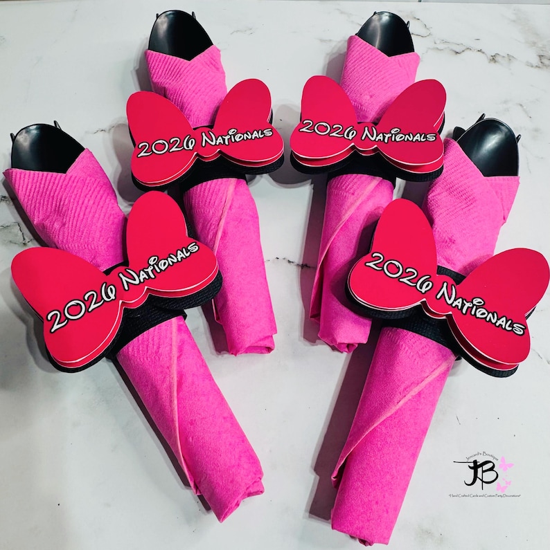 Op de afbeelding: Vier roze servetarrangementen met zwarte keukengerei en rode strikaccenten. Elke strik heeft de tekst "2026 Nationals" in het wit. De servetten zijn opgerold en op een decoratieve manier gerangschikt.