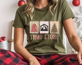 May include: Olive green t-shirt featuring three square designs with Christmas themes. The designs include a Christmas tree, a nativity scene, and another Christmas tree. The words "TRUE STORY" are printed below the designs in a script font.