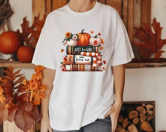 May include: White t-shirt featuring a fall-themed graphic. The design includes a stack of books, pumpkins, flowers, and the text "Just a Girl who loves Fall." The shirt has short sleeves and a relaxed fit, perfect for autumn.