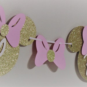 May include: A pink and gold glitter Minnie Mouse themed banner with the letter "B" in the center. The banner is strung together with white string.