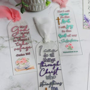 May include: Clear acrylic bookmarks with inspirational quotes and floral accents. One bookmark reads "My God is my rock in whom I take refuge - Psalm 18:2." Another says "I can do all things through Christ who strengthens me - Philippians 4:13."