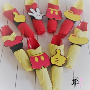 May include: Party decorations featuring red and yellow napkins with red spoons. Each napkin is adorned with a Mickey Mouse-themed cutout, including gloves, shorts, and ears. The decorations are arranged on a white wooden surface.