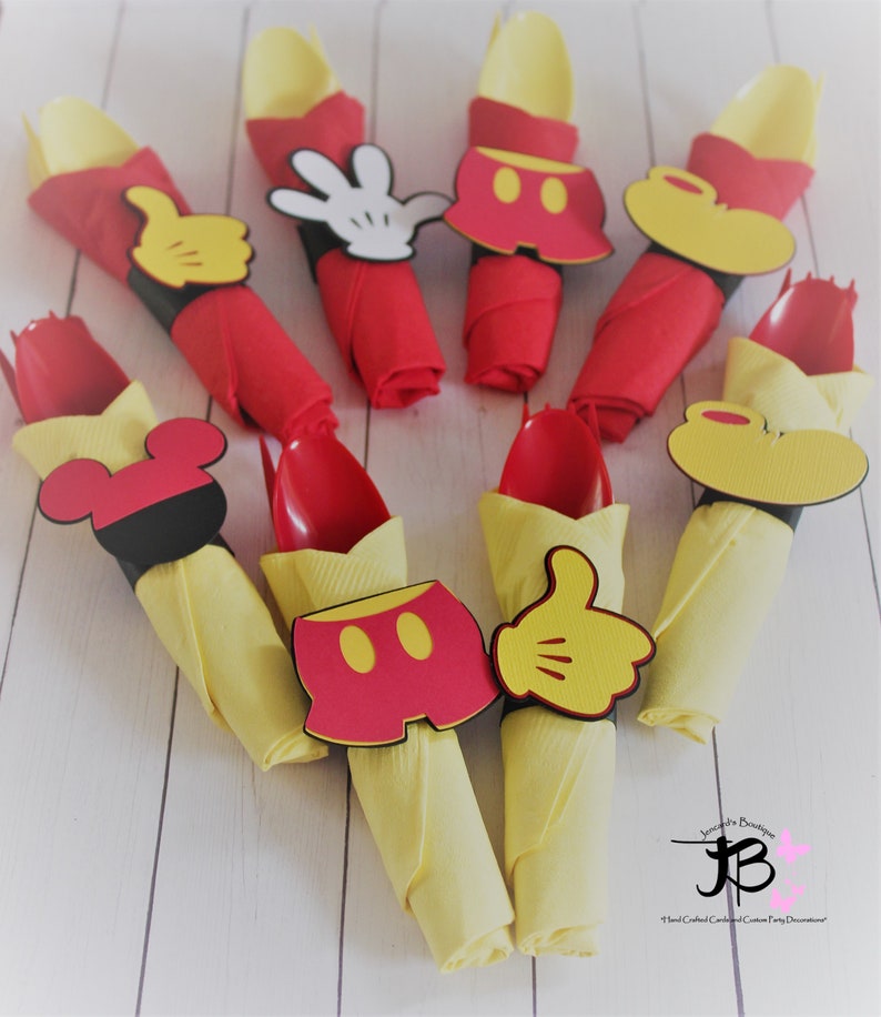May include: Party decorations featuring red and yellow napkins with red spoons. Each napkin is adorned with a Mickey Mouse-themed cutout, including gloves, shorts, and ears. The decorations are arranged on a white wooden surface.