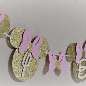 May include: A pink and gold glitter Minnie Mouse themed banner with the letters "D-I-S-N-E-Y" and "B-I" on the circles. The banner is perfect for a birthday party.