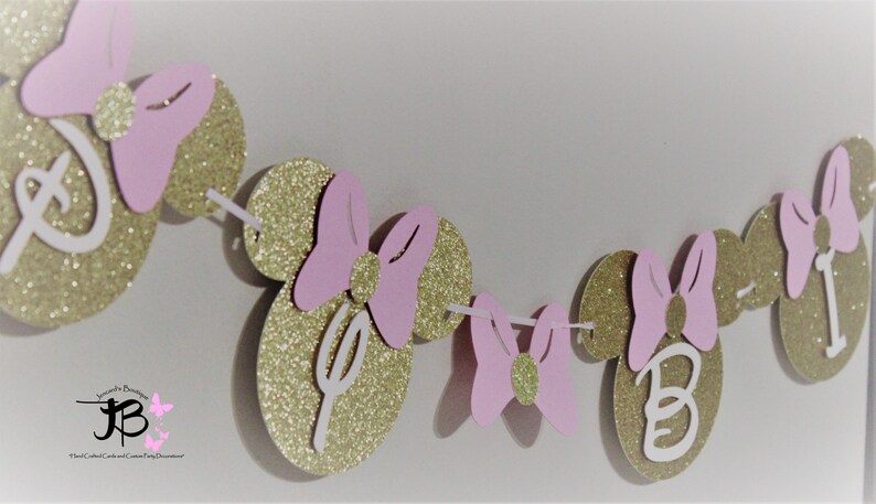 May include: A pink and gold glitter Minnie Mouse themed banner with the letters "D-I-S-N-E-Y" and "B-I" on the circles. The banner is perfect for a birthday party.