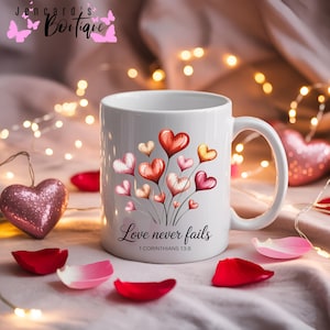 Puede incluir: Taza de cerámica blanca con un diseño de corazones de acuarela en rojo, rosa y amarillo. La taza tiene el texto "Love never fails" y el versículo bíblico 1 Corintios 13:8. Pétalos de rosa y un corazón con purpurina en el fondo.