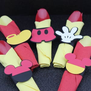 May include: A collection of Mickey Mouse-themed party decorations. The image features red spoons, yellow napkins, and various Mickey Mouse-shaped cutouts, including gloves, shorts, and ears. The decorations are arranged on a dark surface.