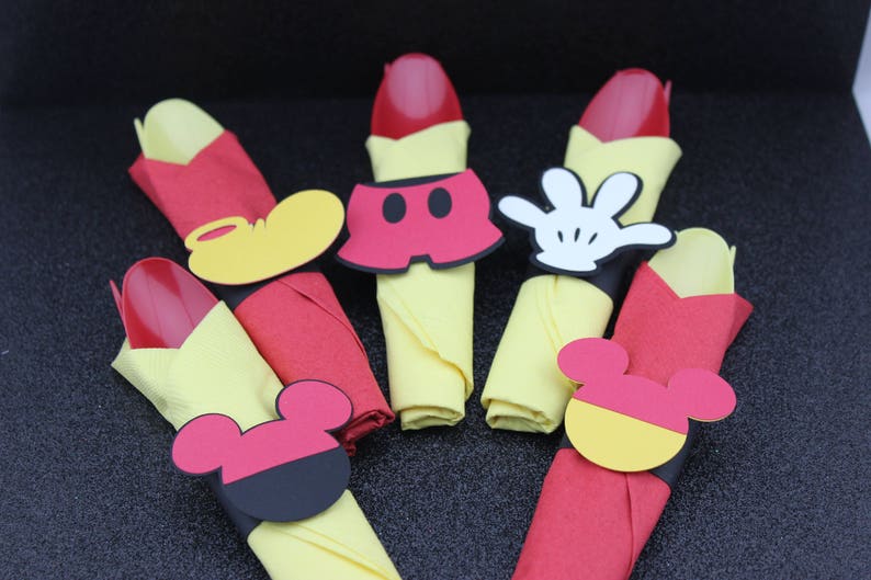 May include: A collection of Mickey Mouse-themed party decorations. The image features red spoons, yellow napkins, and various Mickey Mouse-shaped cutouts, including gloves, shorts, and ears. The decorations are arranged on a dark surface.