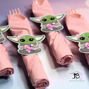 Puede incluir: Cuatro servilletas rosas con un personaje verde que sostiene flores en cada una. El personaje es una versi&oacute;n de dibujos animados de Baby Yoda de la serie Star Wars.