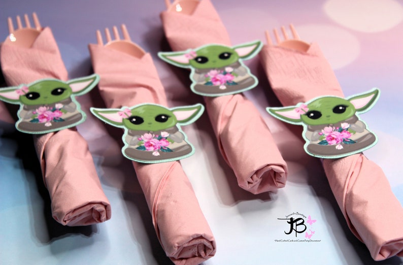 Puede incluir: Cuatro servilletas rosas con un personaje verde que sostiene flores en cada una. El personaje es una versi&oacute;n de dibujos animados de Baby Yoda de la serie Star Wars.