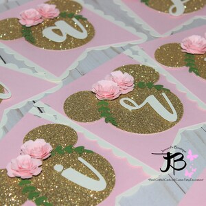 Puede incluir: Bandera de brillo rosa y oro con letras blancas que deletrean "Happy" y un dise&ntilde;o floral. La bandera est&aacute; decorada con flores de papel rosa y hojas verdes.