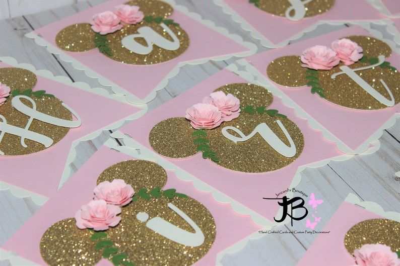 Puede incluir: Bandera de brillo rosa y oro con letras blancas que deletrean "Happy" y un dise&ntilde;o floral. La bandera est&aacute; decorada con flores de papel rosa y hojas verdes.