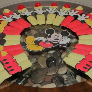 May include: A festive arrangement featuring red and yellow paper napkins, arranged in a circular pattern. Mickey Mouse cutouts and red plastic forks are also included. The centerpiece is set on a glass surface with decorative stones.