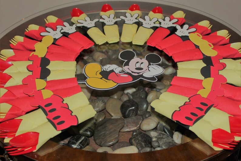 May include: A festive arrangement featuring red and yellow paper napkins, arranged in a circular pattern. Mickey Mouse cutouts and red plastic forks are also included. The centerpiece is set on a glass surface with decorative stones.
