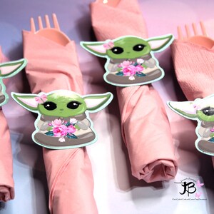 Puede incluir: Cuatro servilletas rosas con un personaje verde sosteniendo flores impresas en ellas. El personaje lleva una t&uacute;nica marr&oacute;n y tiene ojos grandes. Las servilletas est&aacute;n enrolladas y atadas con un tenedor.