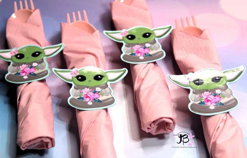 Puede incluir: Cuatro servilletas rosas con un personaje verde sosteniendo flores impresas en ellas. El personaje lleva una t&uacute;nica marr&oacute;n y tiene ojos grandes. Las servilletas est&aacute;n enrolladas y atadas con un tenedor.
