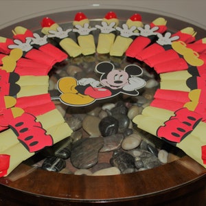 May include: A decorative table centerpiece featuring Mickey Mouse. The arrangement includes red and yellow paper cutouts resembling Mickey's gloves and shorts, arranged around a central Mickey Mouse image. The table is made of glass and has a wooden base.