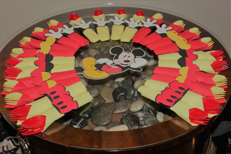 May include: A decorative table centerpiece featuring Mickey Mouse. The arrangement includes red and yellow paper cutouts resembling Mickey's gloves and shorts, arranged around a central Mickey Mouse image. The table is made of glass and has a wooden base.