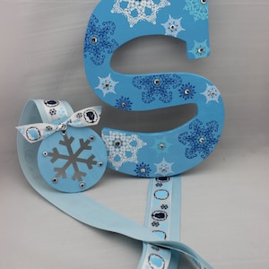 May include: A light blue wooden letter S decorated with white and blue snowflake designs and rhinestones. A blue ribbon with a white snowflake ornament is attached to the letter.