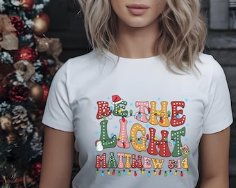 Peut inclure: T-shirt blanc avec l'inscription "BE THE LIGHT MATTHEW 5:14" dans une typographie colorée et festive. Le motif comprend des guirlandes lumineuses et un bonnet de Père Noël, suggérant un thème de vacances.