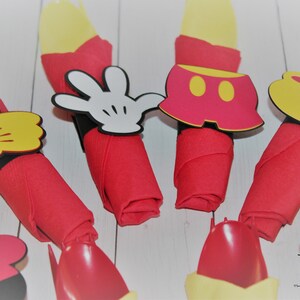 May include: Red cloth napkins with yellow utensils are decorated with Mickey Mouse-themed cutouts. The cutouts include a white glove, yellow thumbs up, red shorts, and a yellow shoe. The napkins are arranged on a white surface.